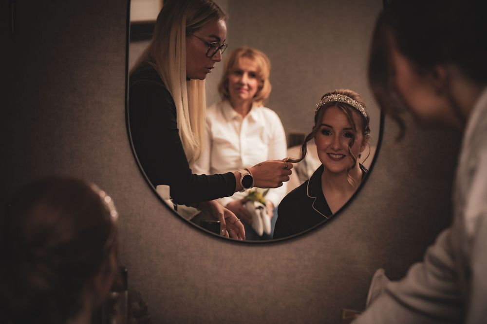 A reflection in a round mirror shows wedding preparations with a white-clad figure getting ready in warm lighting.