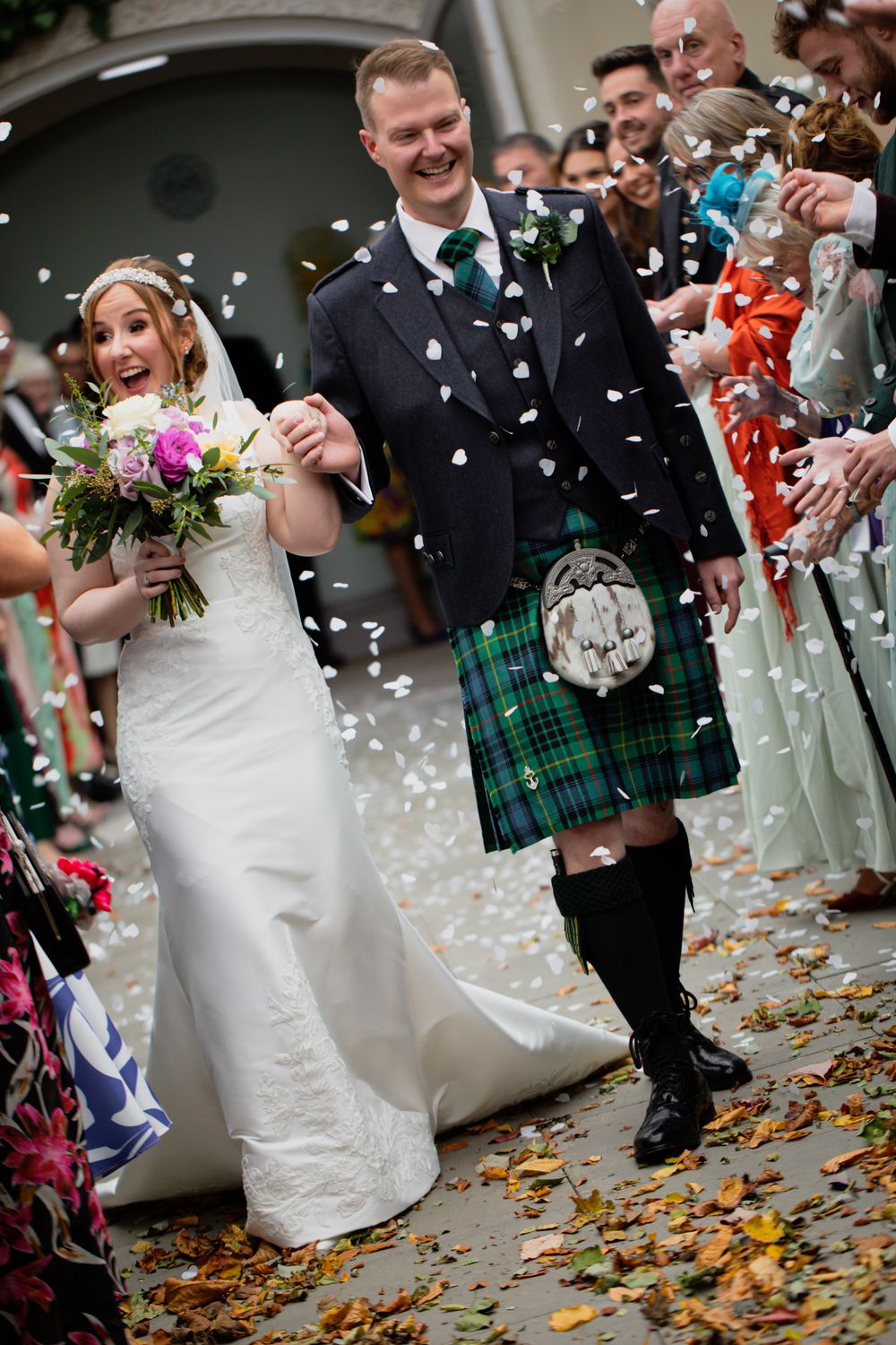 Wedding guests shower the newlyweds with white confetti as they walk together on fallen autumn leaves.