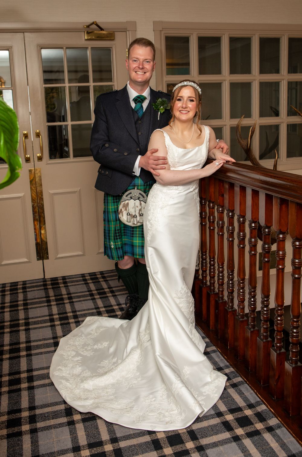 A wedding couple poses on a plaid carpeted staircase, with the groom wearing a traditional kilt and the bride in a white gown.