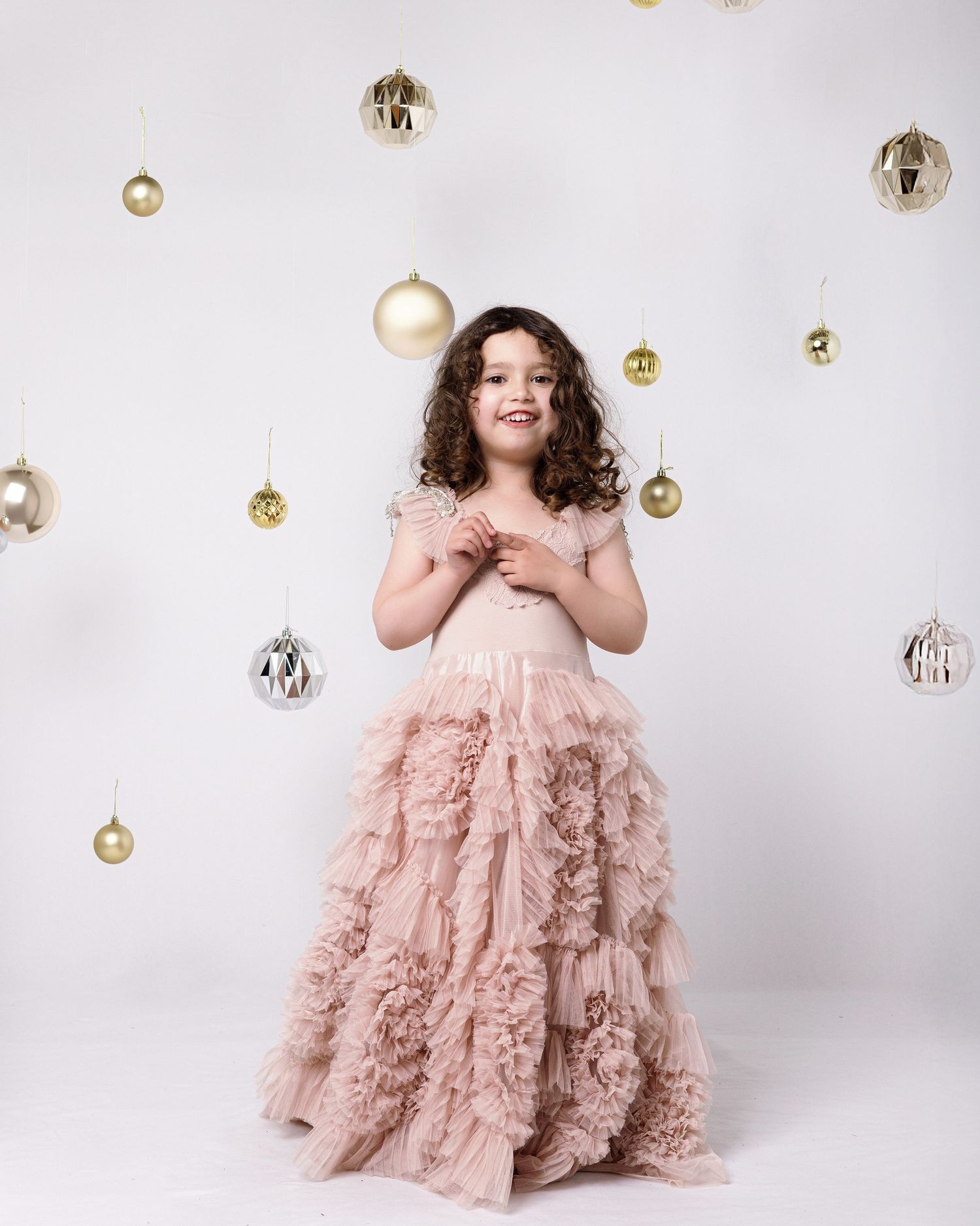 A child in a flowing pink tulle dress poses among hanging gold and silver Christmas ornaments against a white backdrop.
