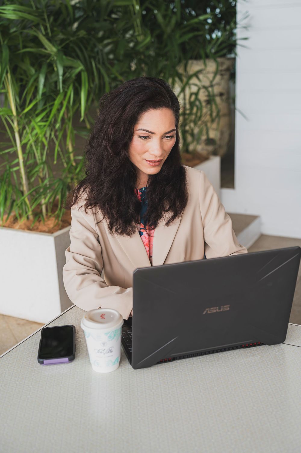 Professional working on laptop at white desk with coffee cup and indoor plants in background.