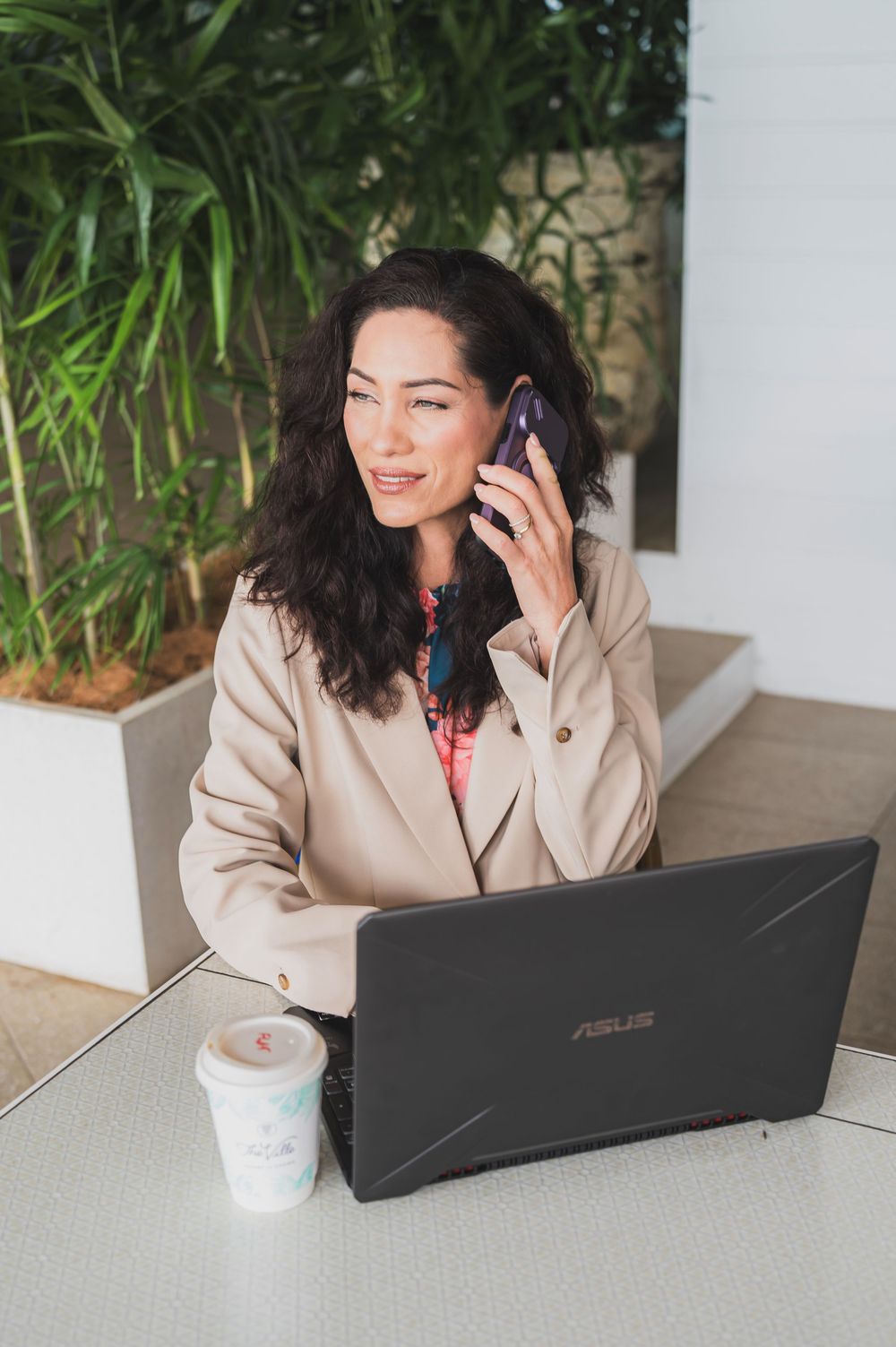 Professional working on Asus laptop at outdoor cafe with phone and coffee, wearing beige coat.