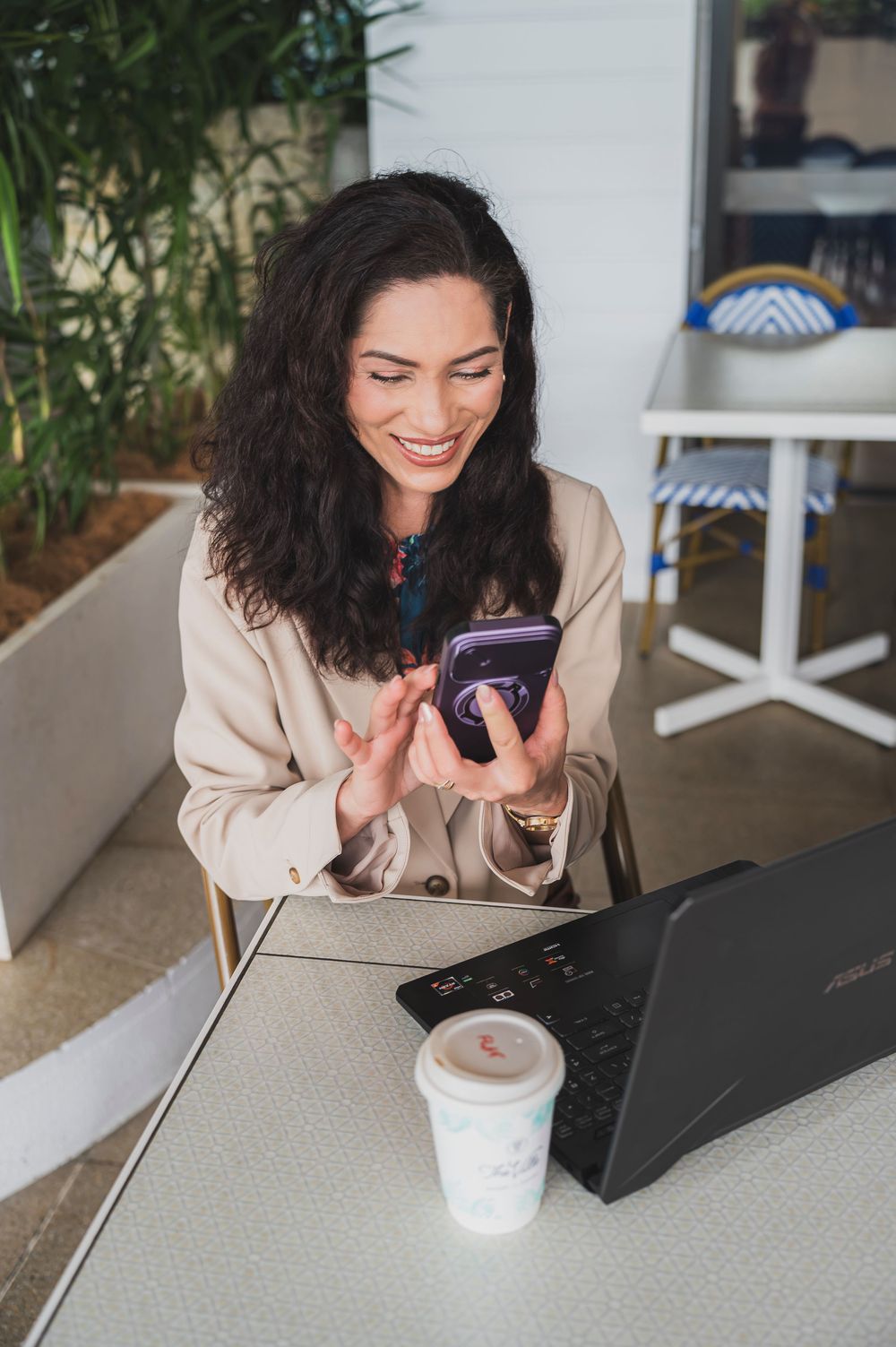 A person with curly dark hair in a beige blazer smiles while using a laptop and phone at a white desk.
