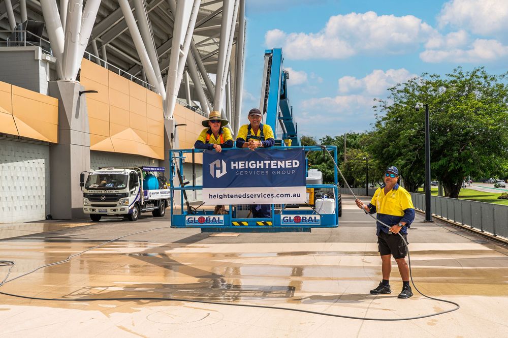 Workers in yellow safety vests operate a pressure washer to clean the exterior of a stadium building on a sunny day.