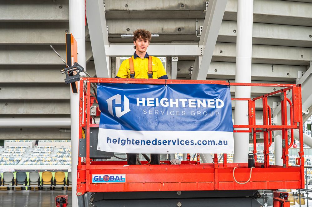 A worker in yellow safety gear operates an elevated red scissor lift platform with a Heightened Services banner.