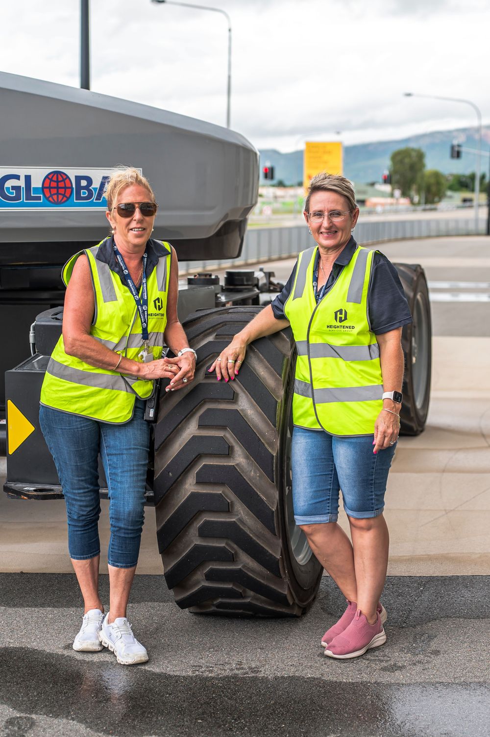 Two workers in safety vests stand next to a large industrial tractor tire at a worksite.