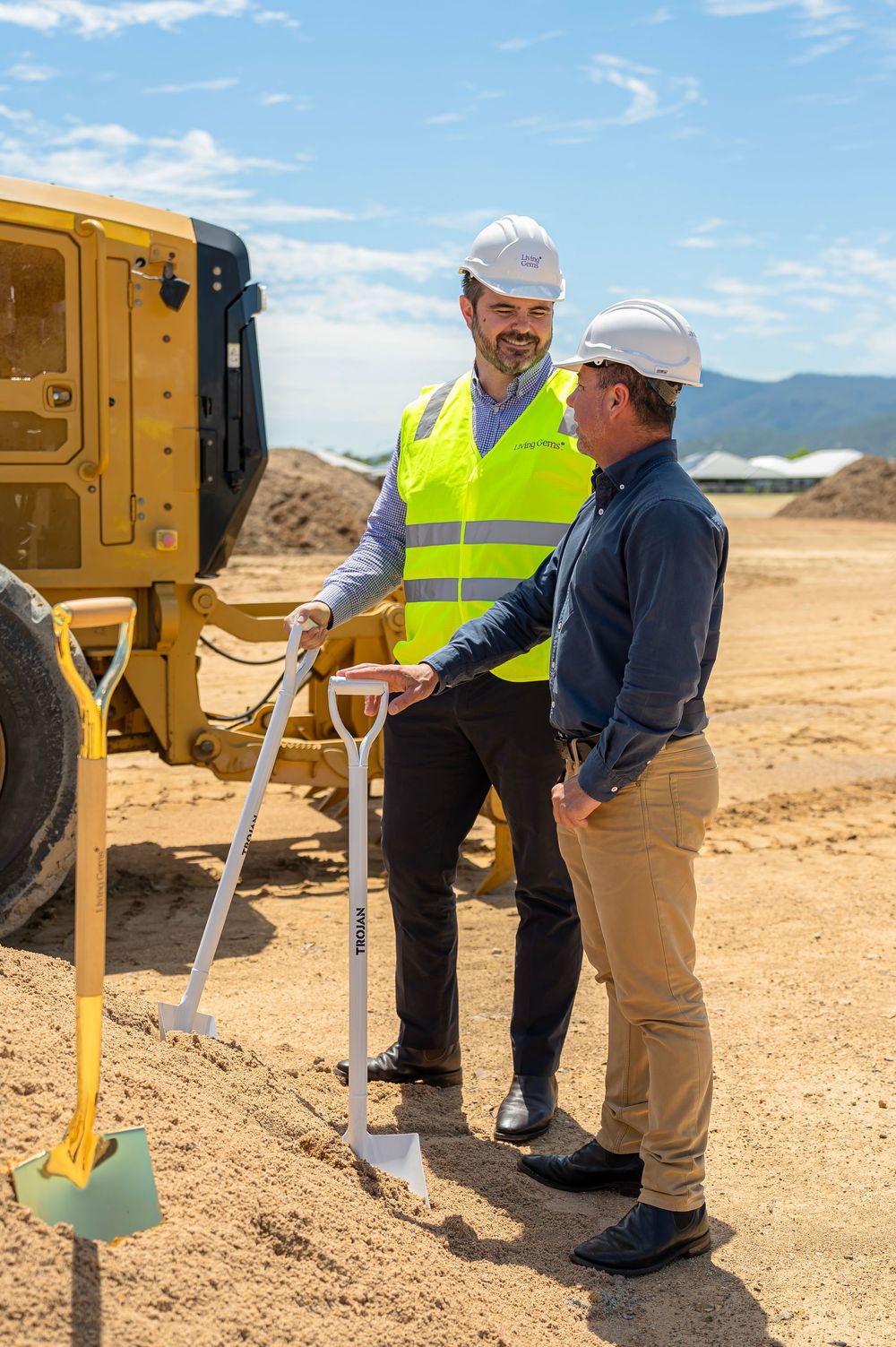 Construction workers in safety gear discuss plans while standing next to heavy equipment at a dusty job site.
