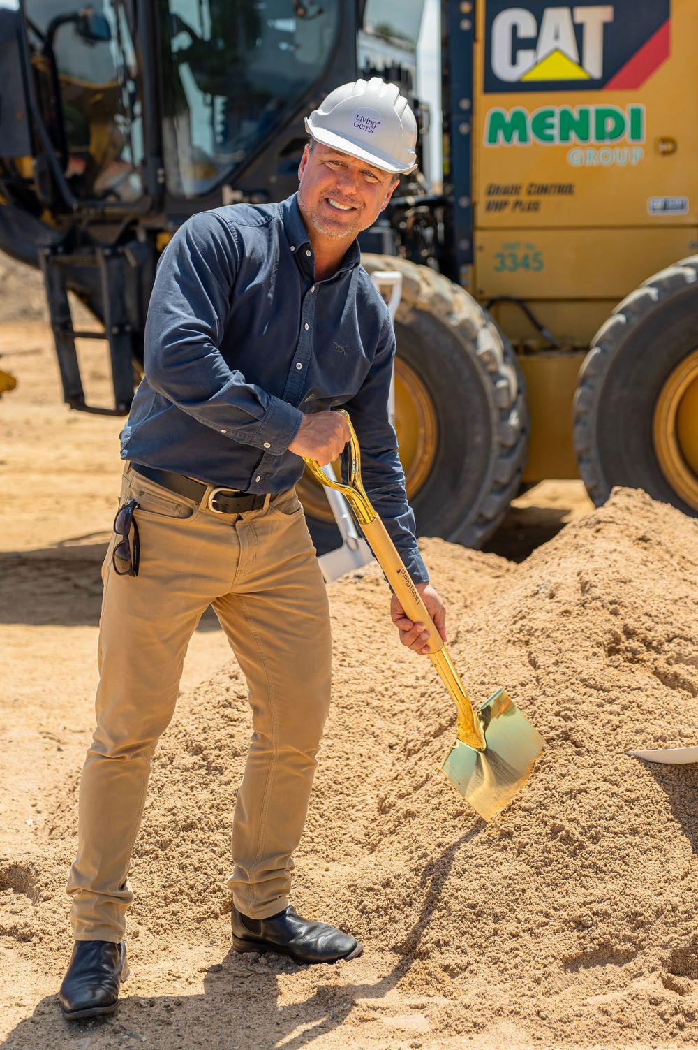 Construction worker in hard hat uses measuring tools on sandy ground near heavy equipment.