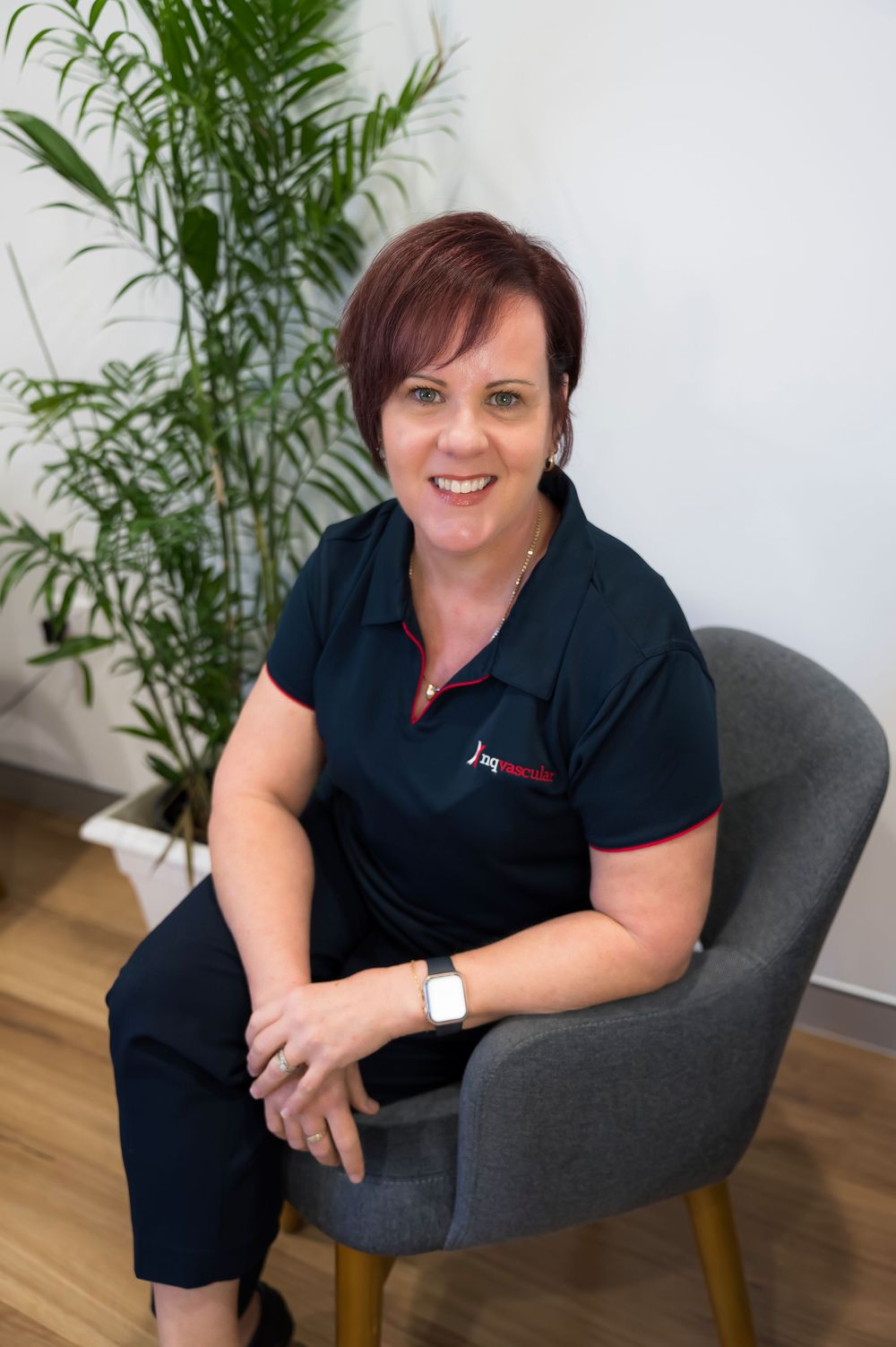 Professional wearing a navy polo shirt sits in a modern grey chair with a decorative palm plant in the background.