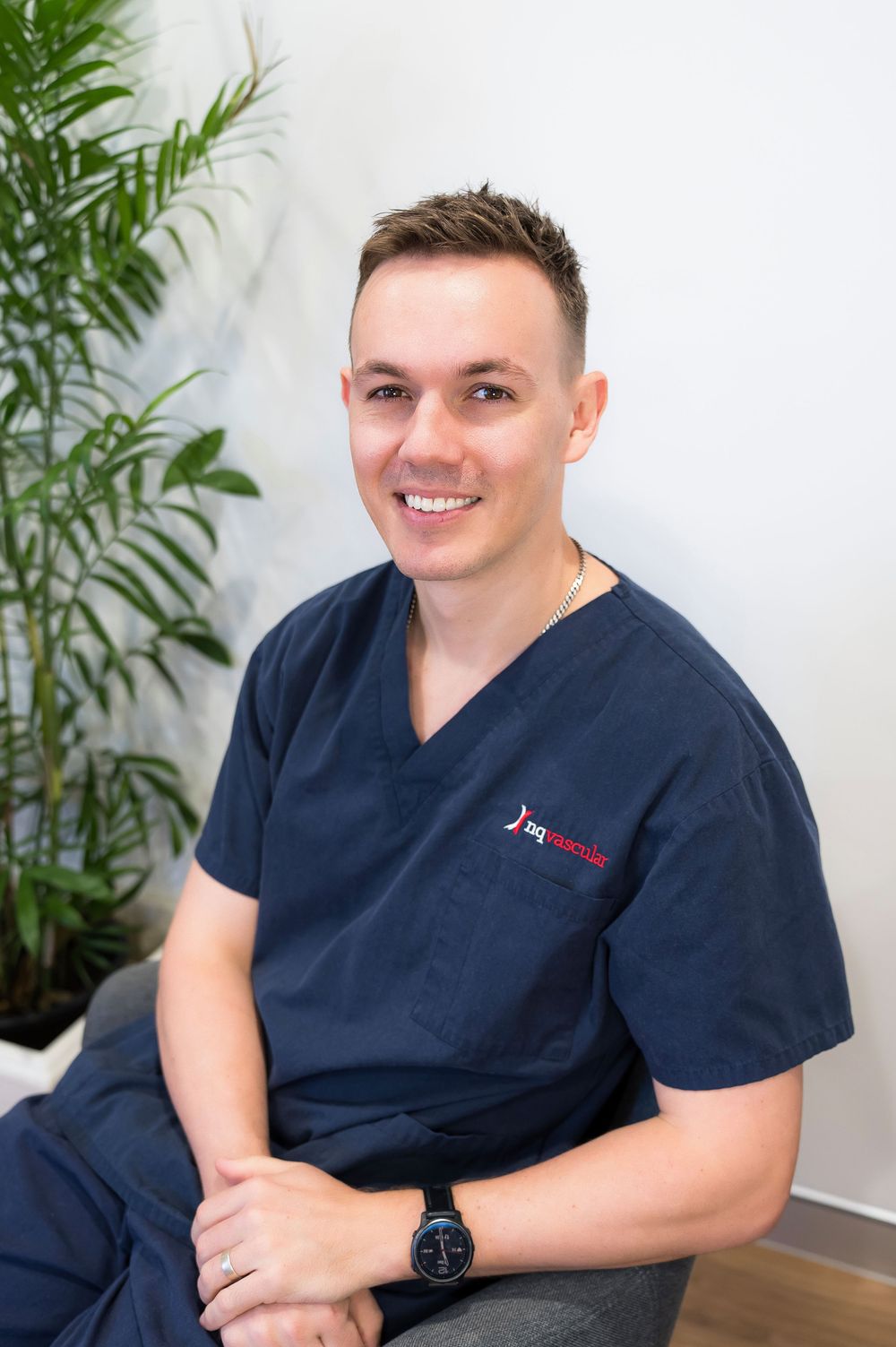 Healthcare professional in navy blue scrubs smiling while seated in a bright modern office setting.