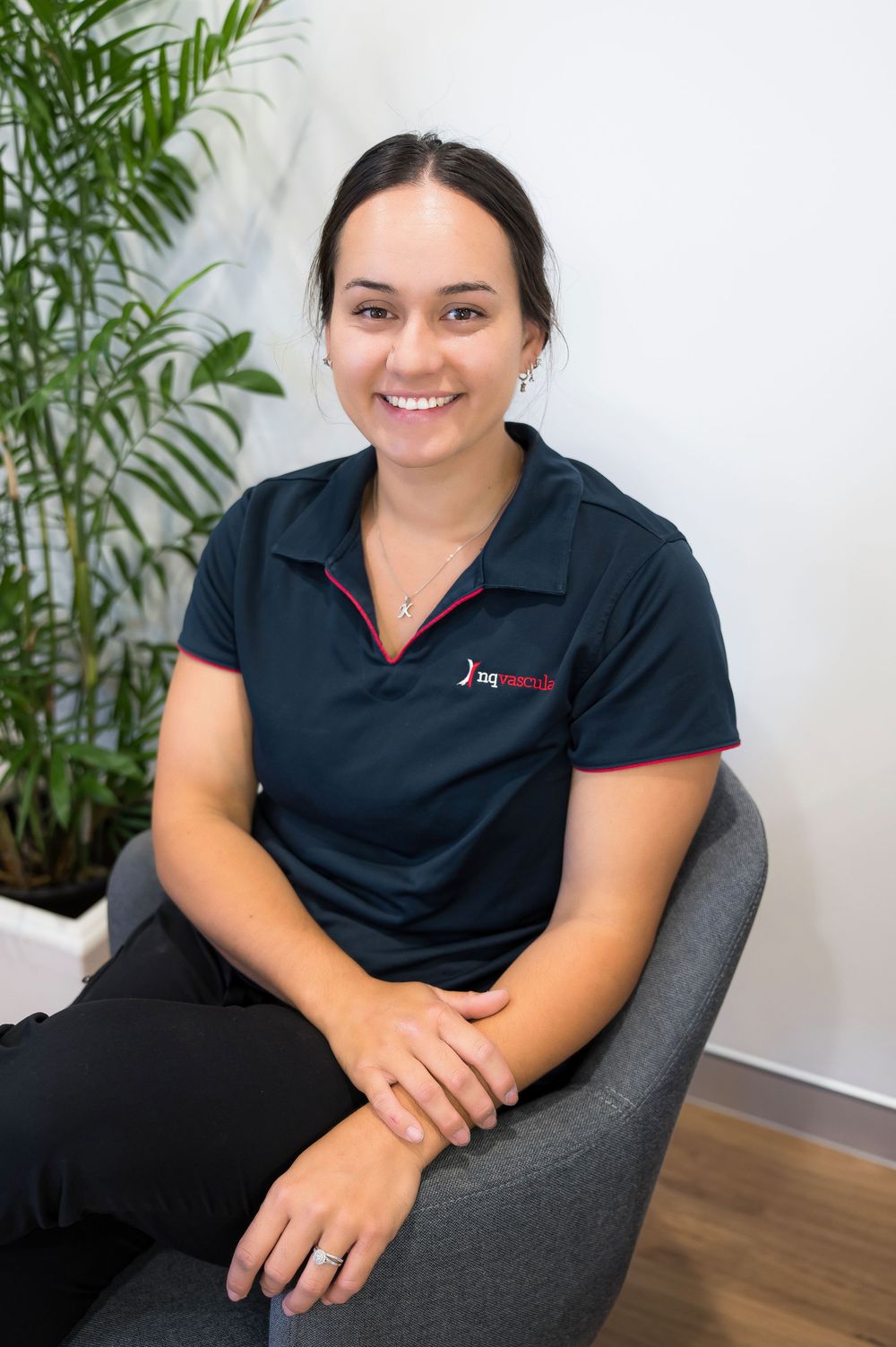 Professional staff member in navy blue polo shirt sitting in gray office chair with palm plant in background.