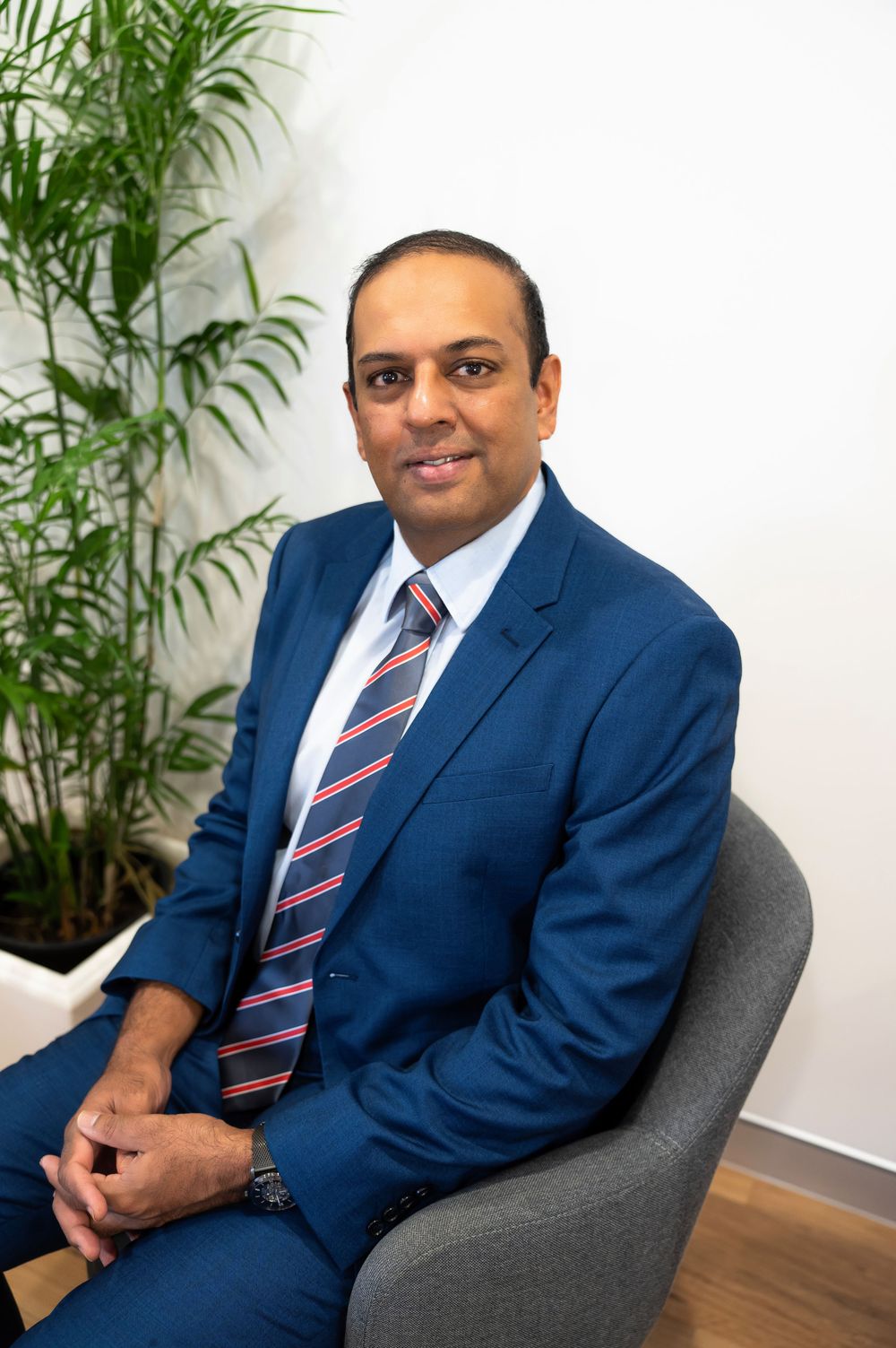 Professional portrait of a person in a navy blue suit and striped tie seated in a gray chair with indoor plants in background.