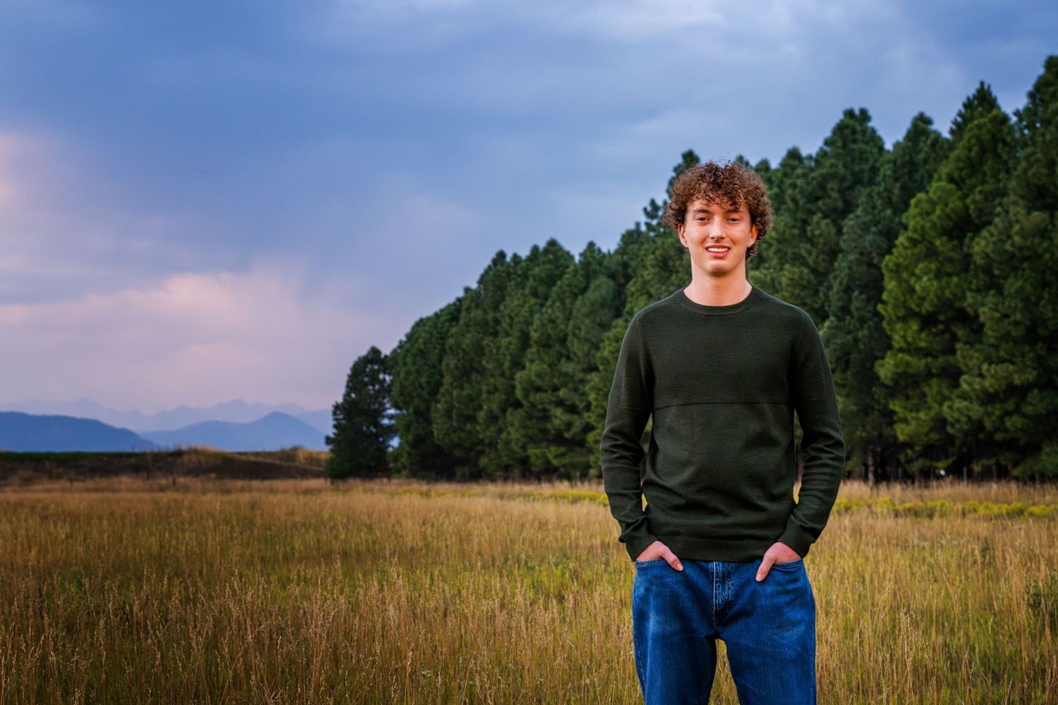 A Denver Senior in a dark sweater stands in a golden field at dusk with evergreen trees and mountains in the background.
