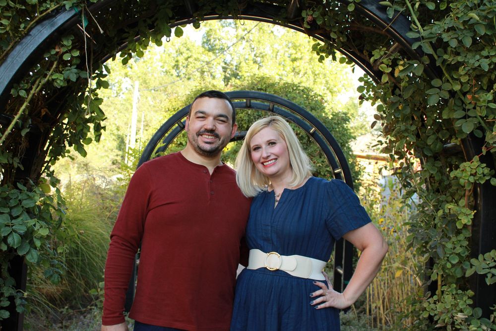 A couple poses outdoors for engagement photos in a natural setting with sunlit foliage in the background.