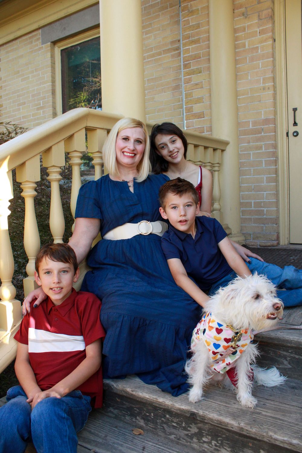 Family group sits on porch steps with small white dog wearing colorful bandana.