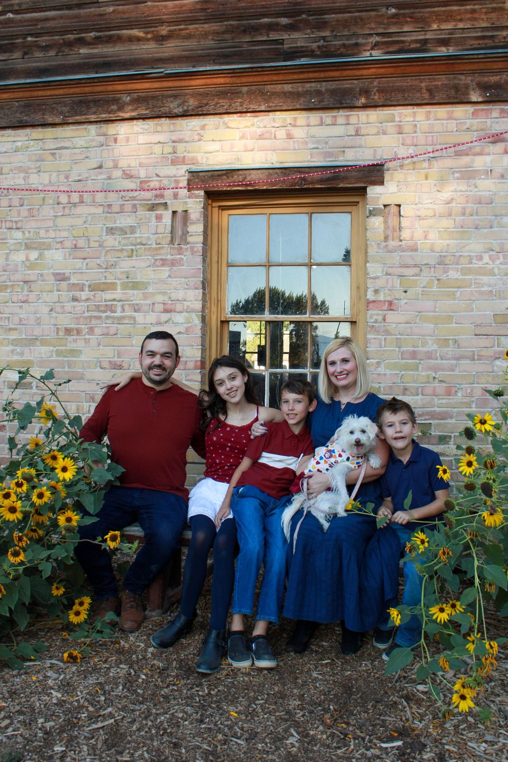 A group sits together on a rustic brick porch surrounded by blooming sunflowers on an autumn day.