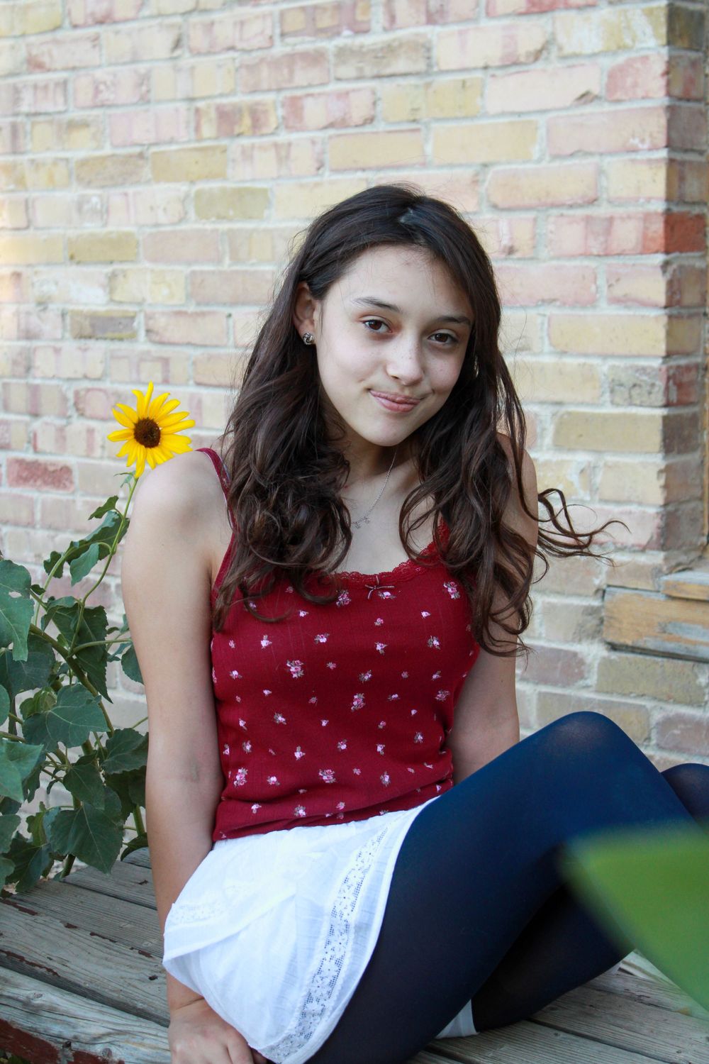 Portrait of young woman in red top sitting on bench with sunflower in background.