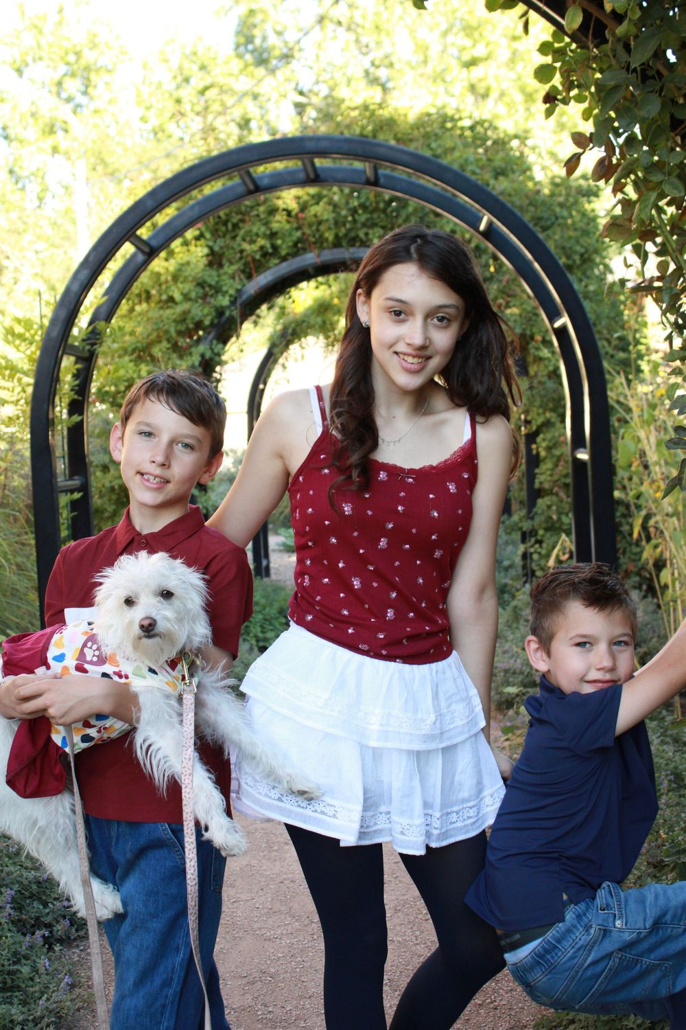 Family poses together with small white dog under garden arch in outdoor portrait setting.