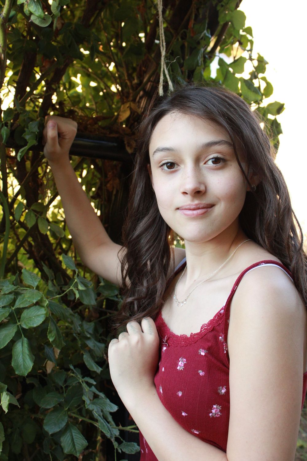 A person in a burgundy top poses in front of lush green foliage on a sunny day.