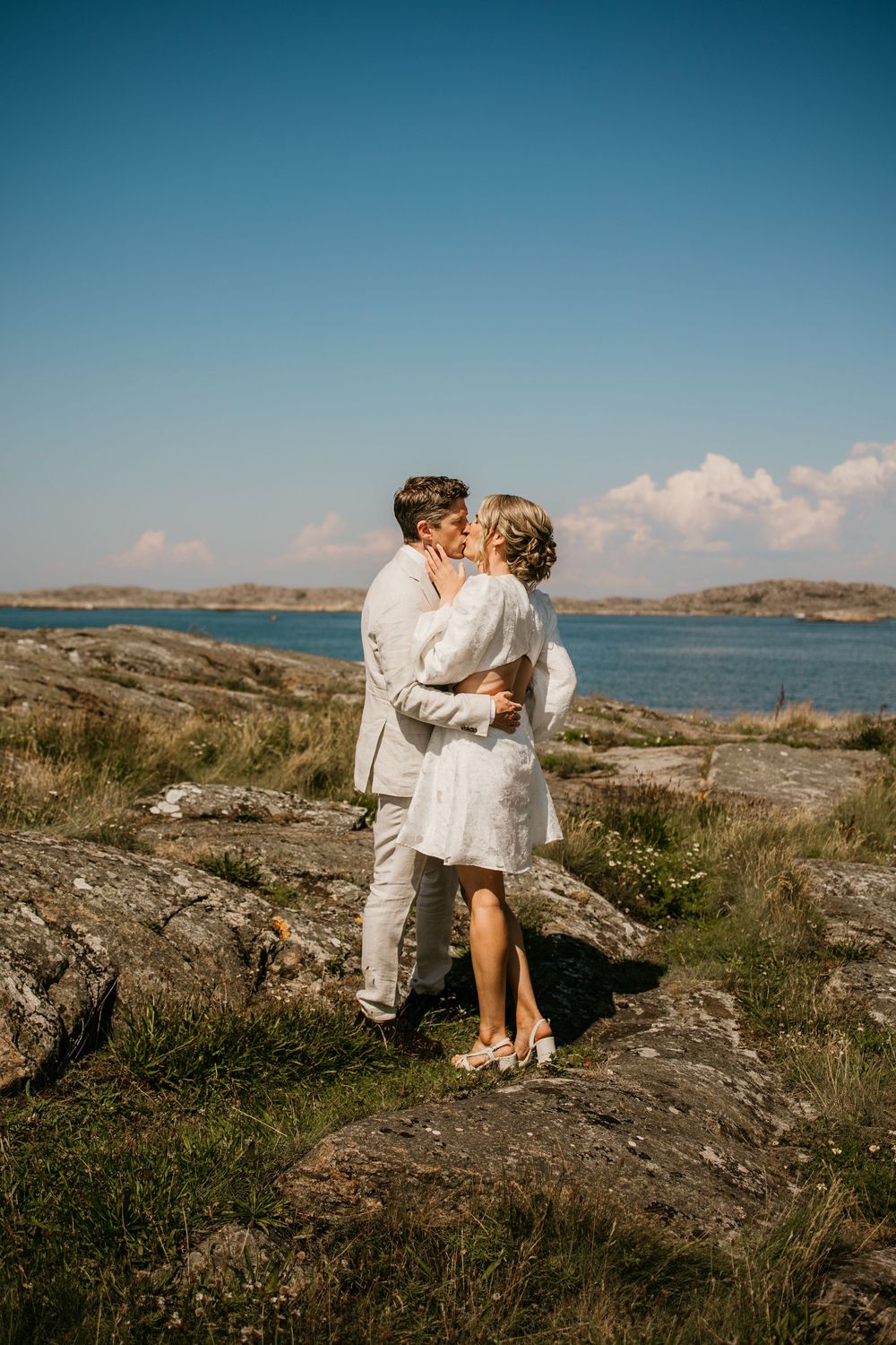 A romantic couple in white attire share intimate moments on rocky coastal cliffs overlooking a serene ocean bay.