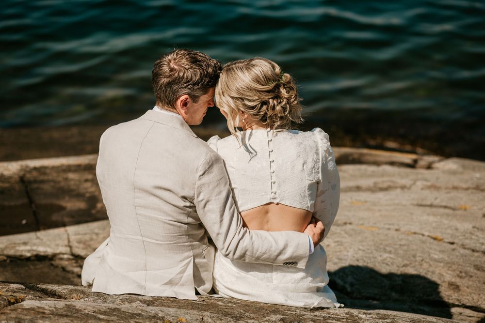 A couple sits together on rocky shoreline overlooking dark rippling water during an intimate outdoor moment.