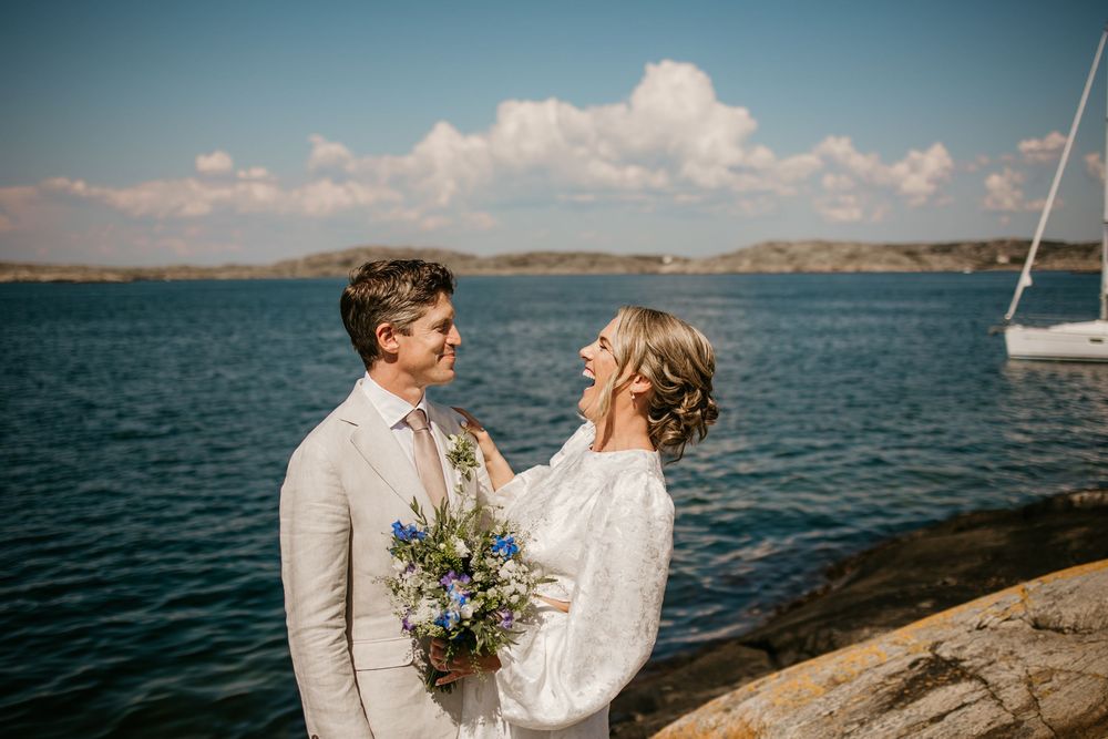 A couple in wedding attire shares a romantic moment on rocky coastline with blue water and sailboat in background.
