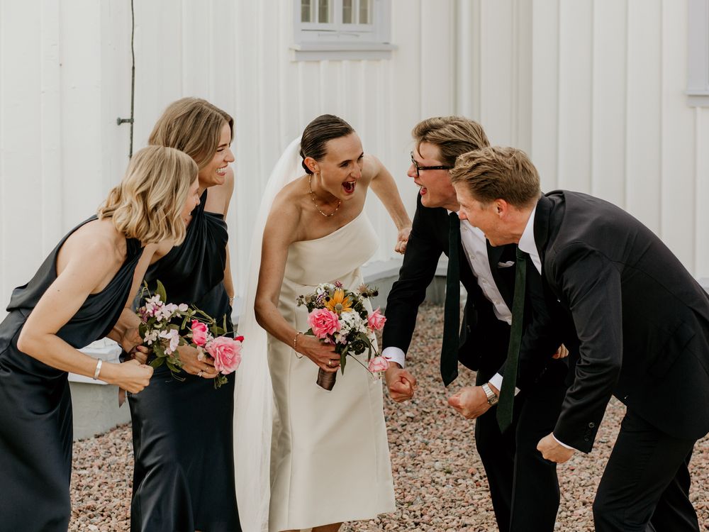 Wedding party members laugh and share a joyful moment together on a gravel path next to a white building.
