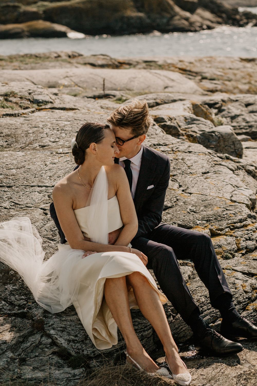 A couple in wedding attire share an intimate moment while sitting on coastal rocks at sunset.