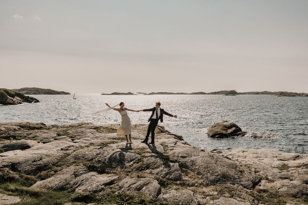 Two figures hold hands while balancing on rocky coastal shoreline at sunset with ocean waves in background.