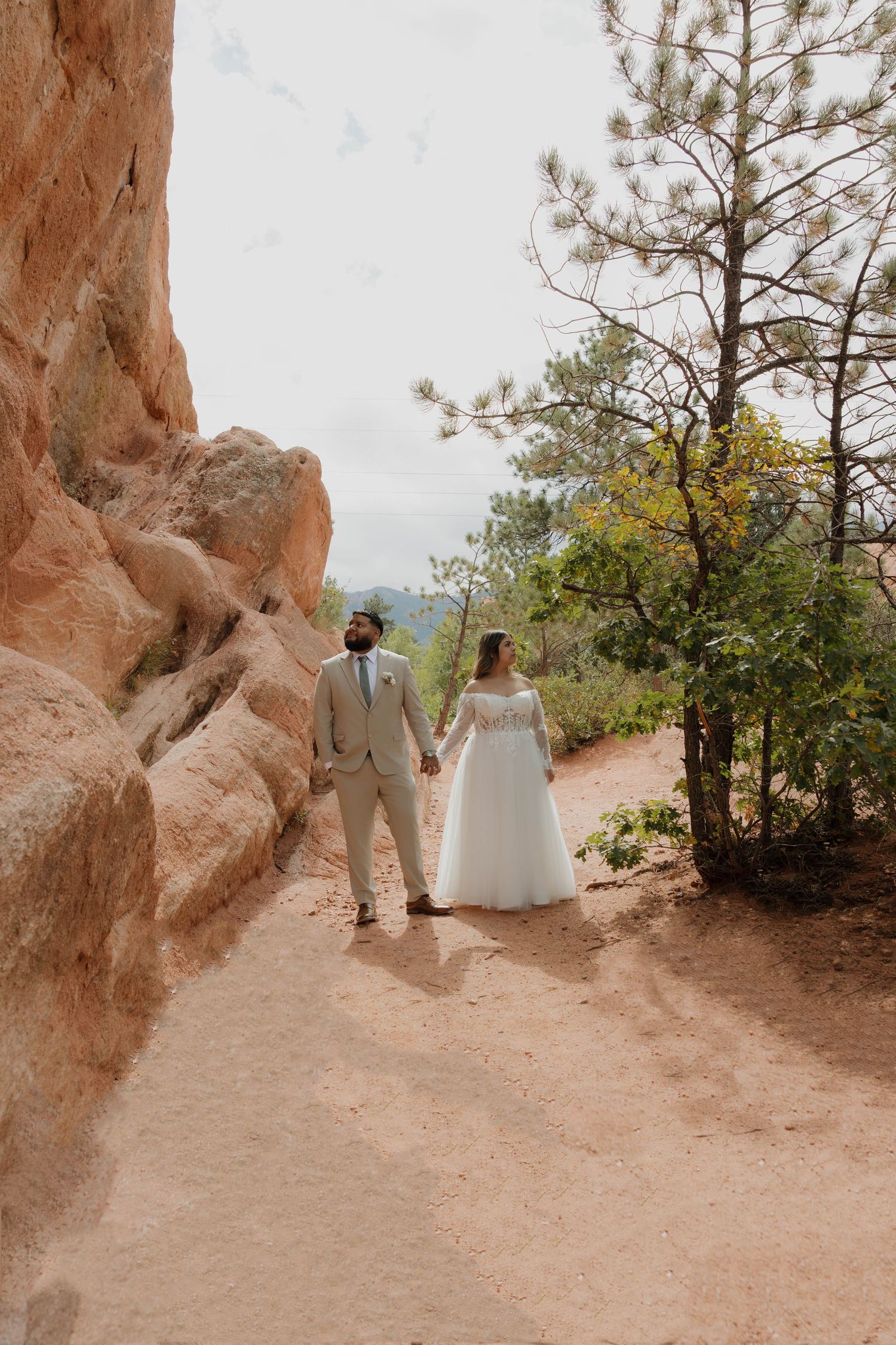 A couple walks along a scenic desert trail surrounded by red rock formations and pine trees in the distance.