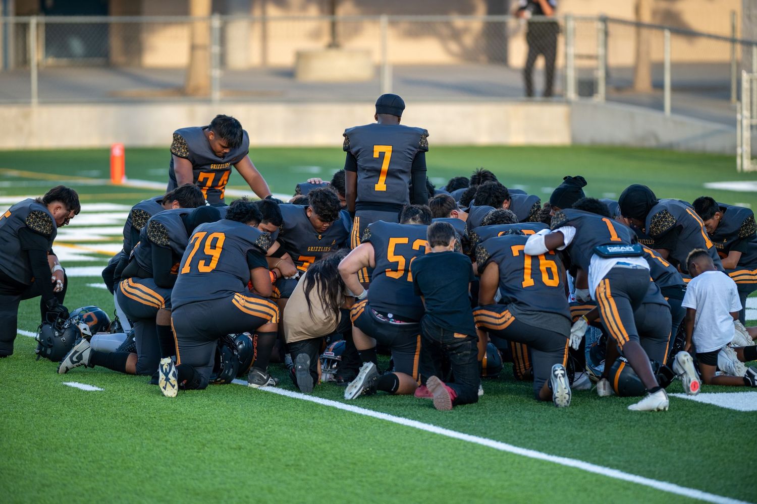 Football team gathers on field sideline during practice or game preparation.