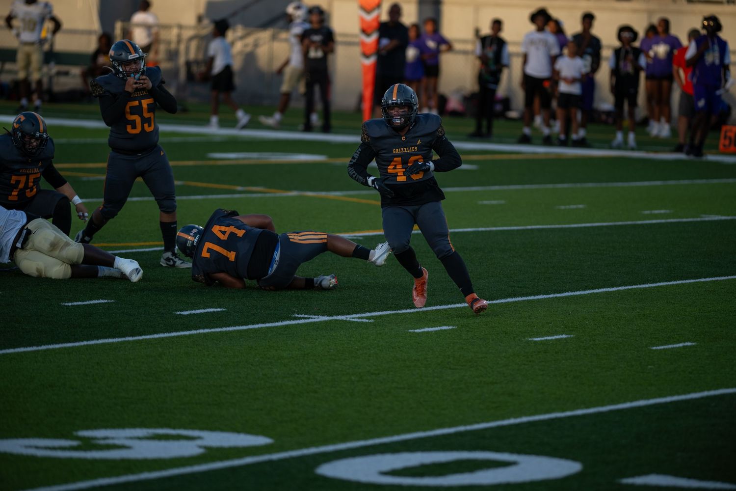 Young football teams face off during a youth league game on an artificial turf field under bright lighting.