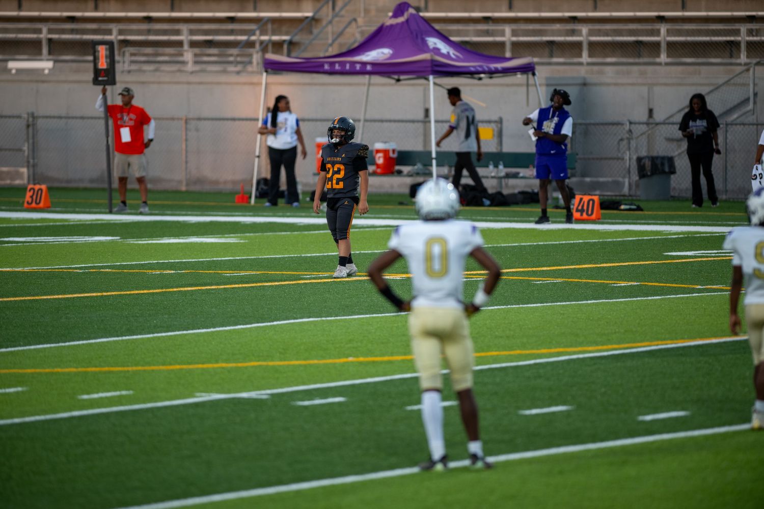 Multiple football players practicing on artificial turf field with goal posts visible in background.