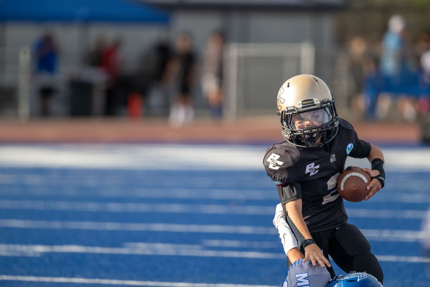 Youth football player in black uniform carrying ball on blue turf field.