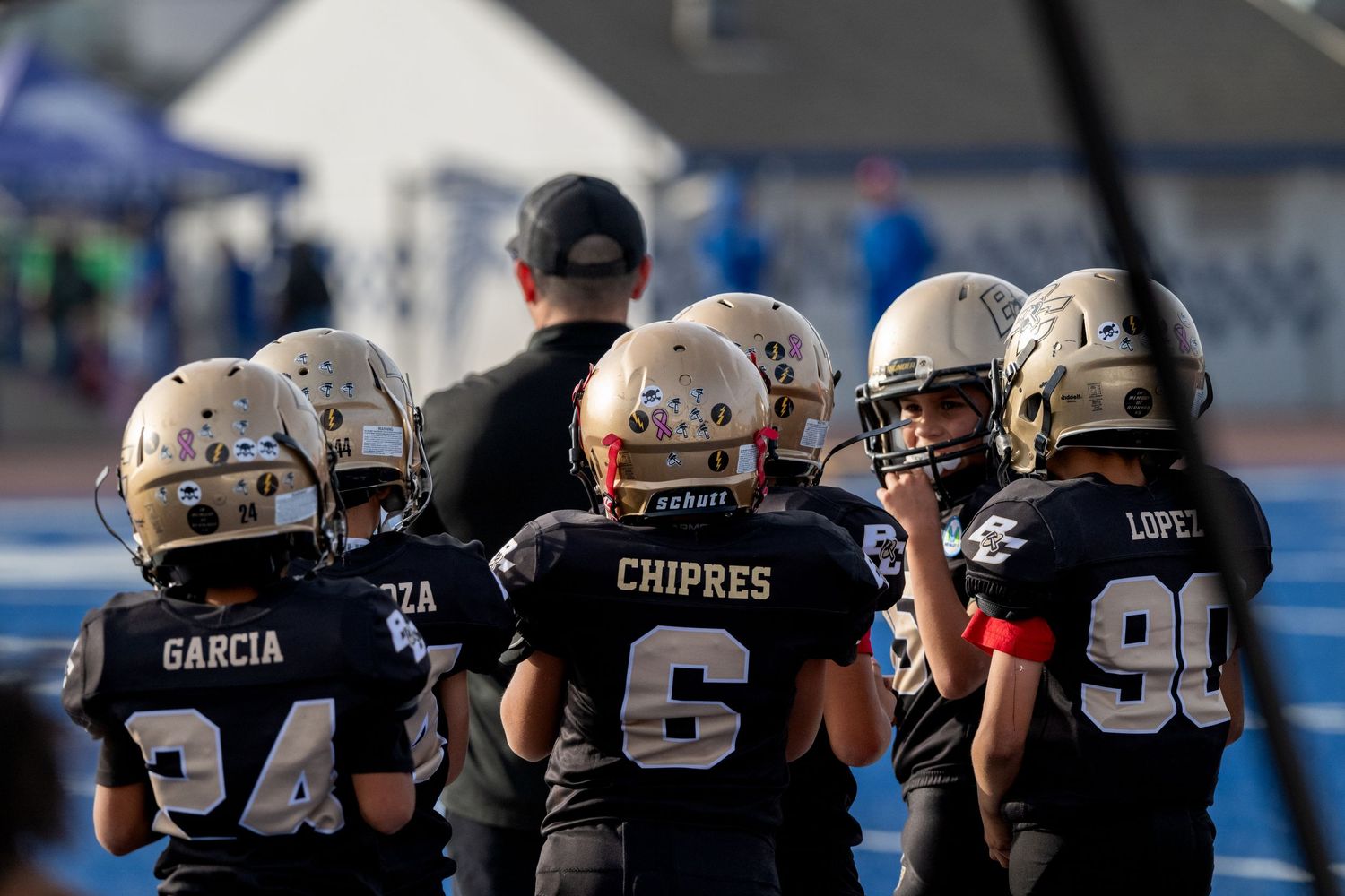 Youth football players in black jerseys huddled on sideline during game.