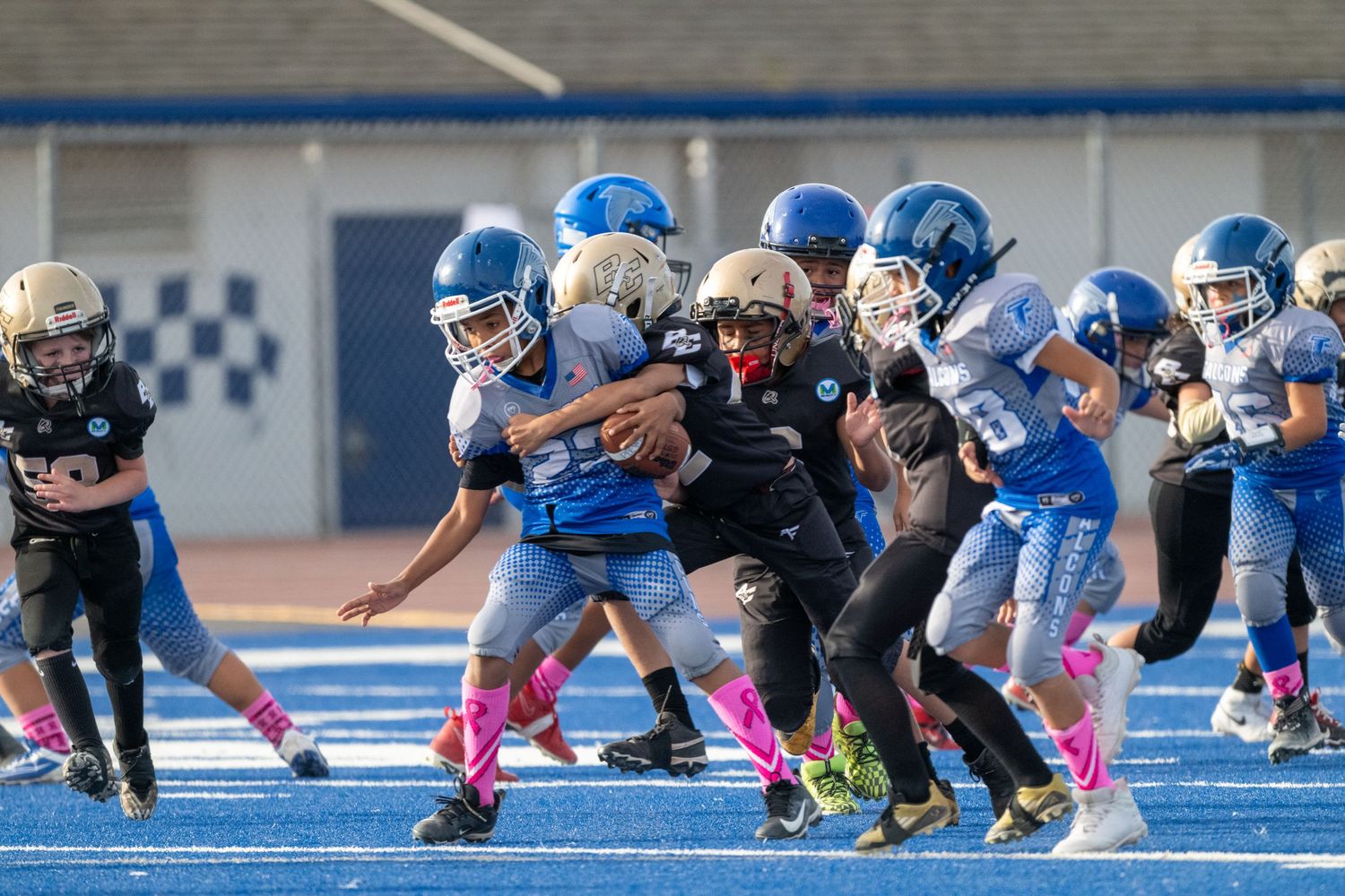 Youth football players in blue and black uniforms battling at line of scrimmage.