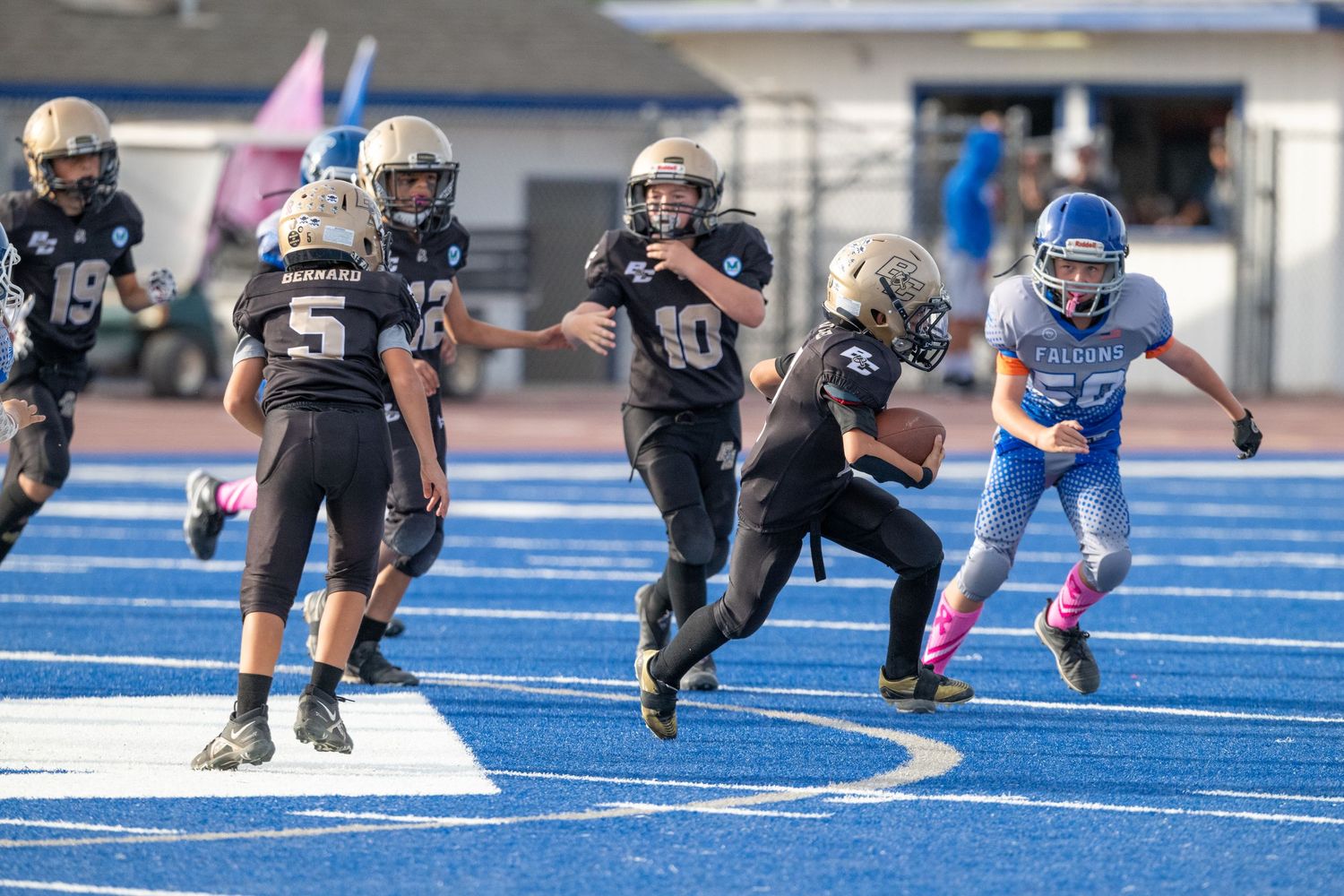 Youth football players in black uniforms running play against blue team on turf field.