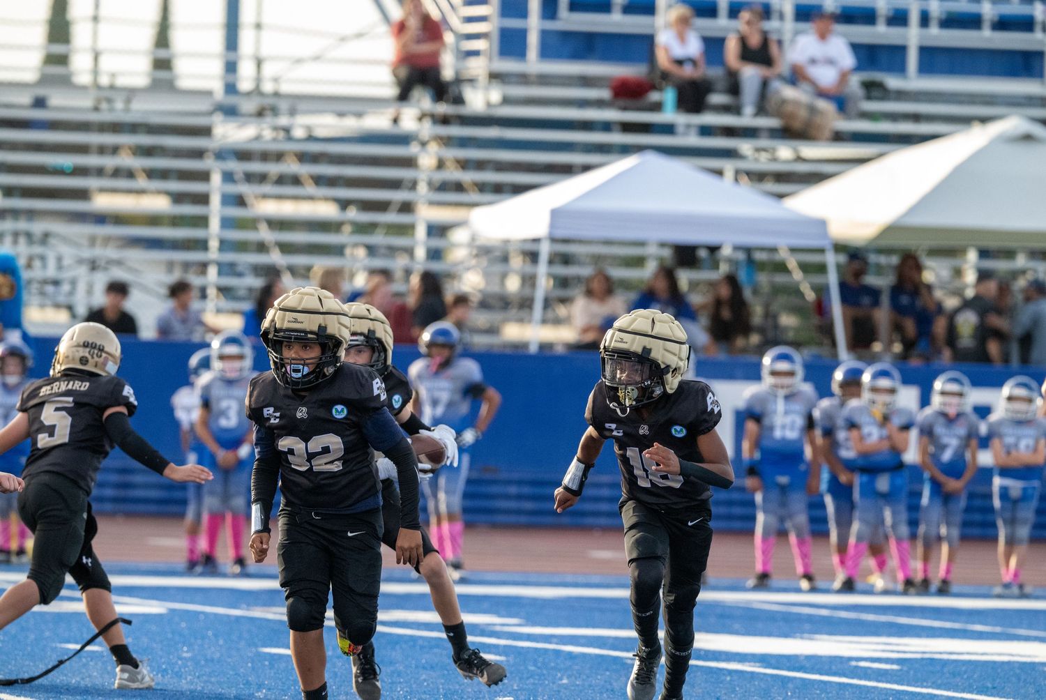 Football scrimmage taking place on distinctive bright blue playing surface with spectators watching.