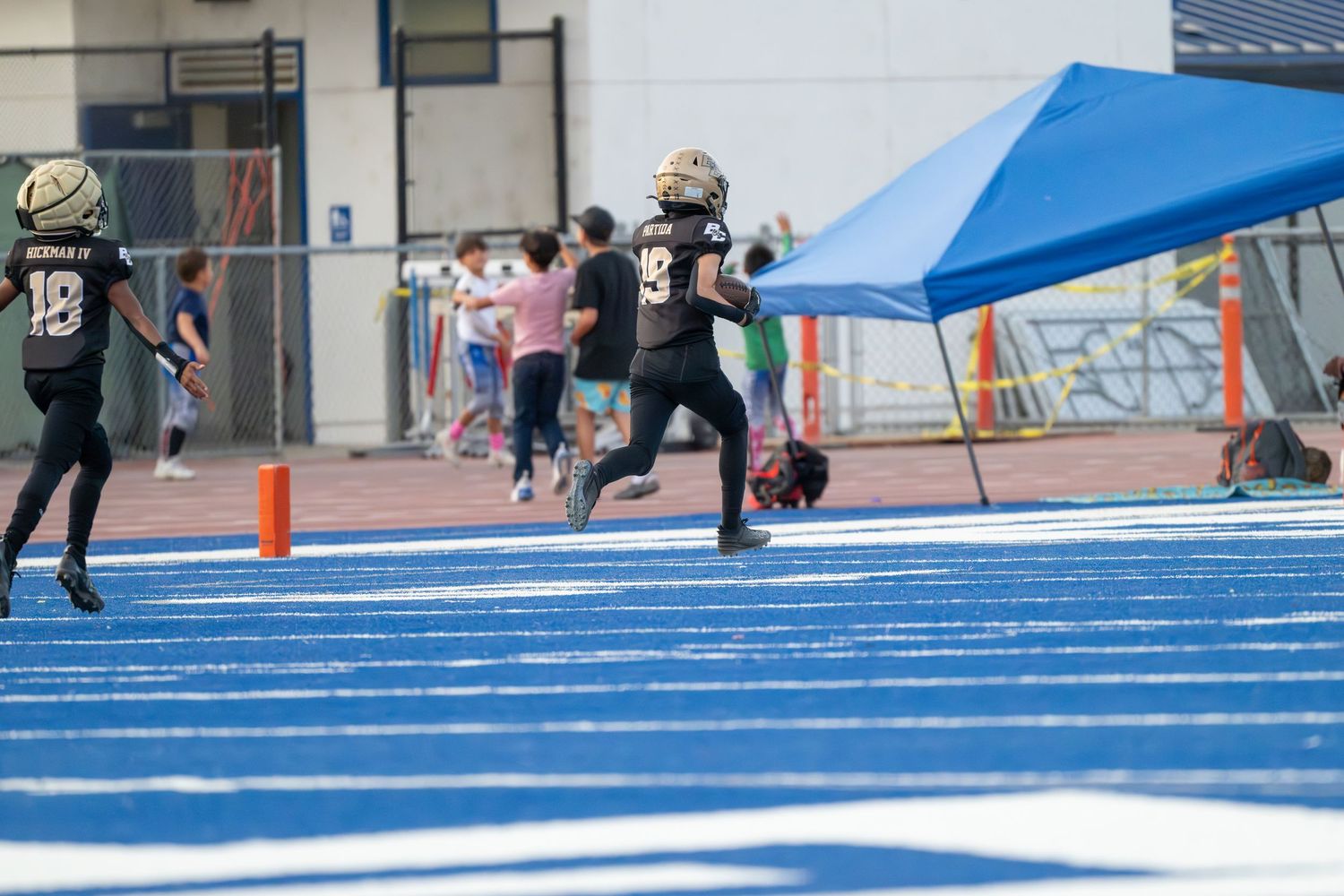 Athletes practicing football drills on blue artificial turf field under sunny skies.