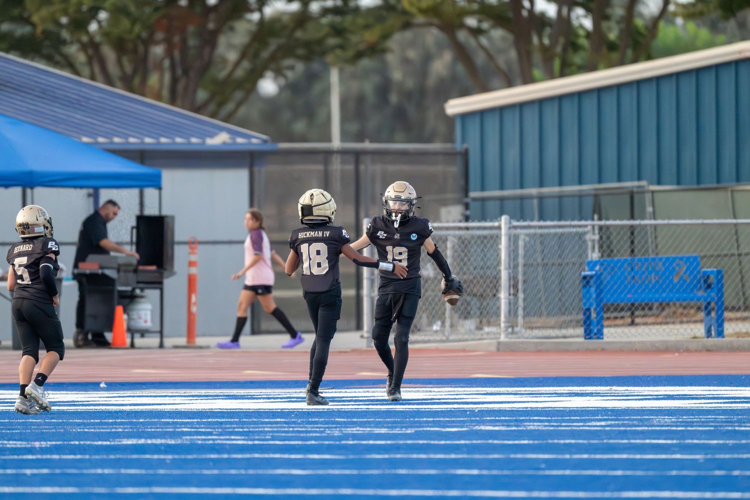Football players running drills on vibrant blue sports field near athletic facilities.
