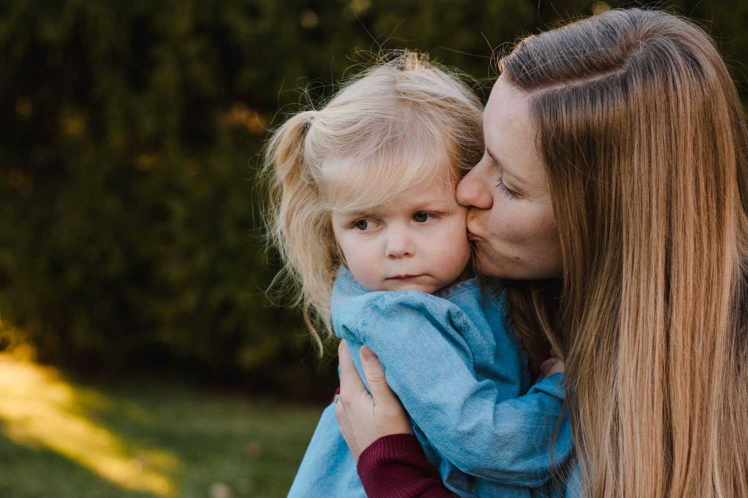 Mother kissing the cheek of her young blonde daughter outdoors in a garden setting.