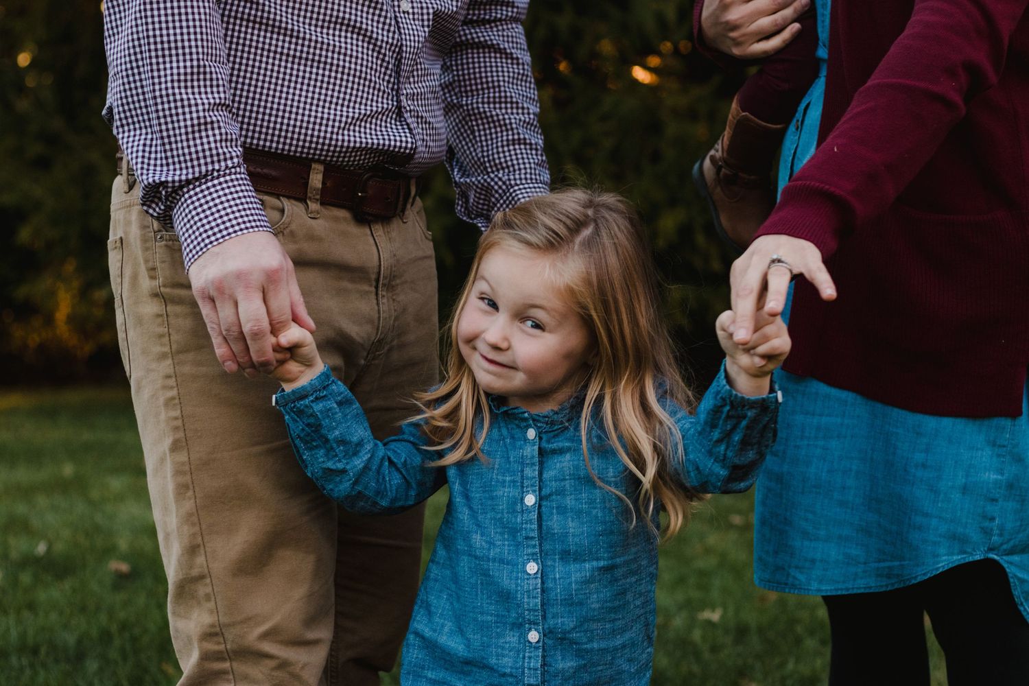 Little girl with goofy smile holding parents hands - South Lyon, Michigan lifestyle family photographer captures joyful moments.