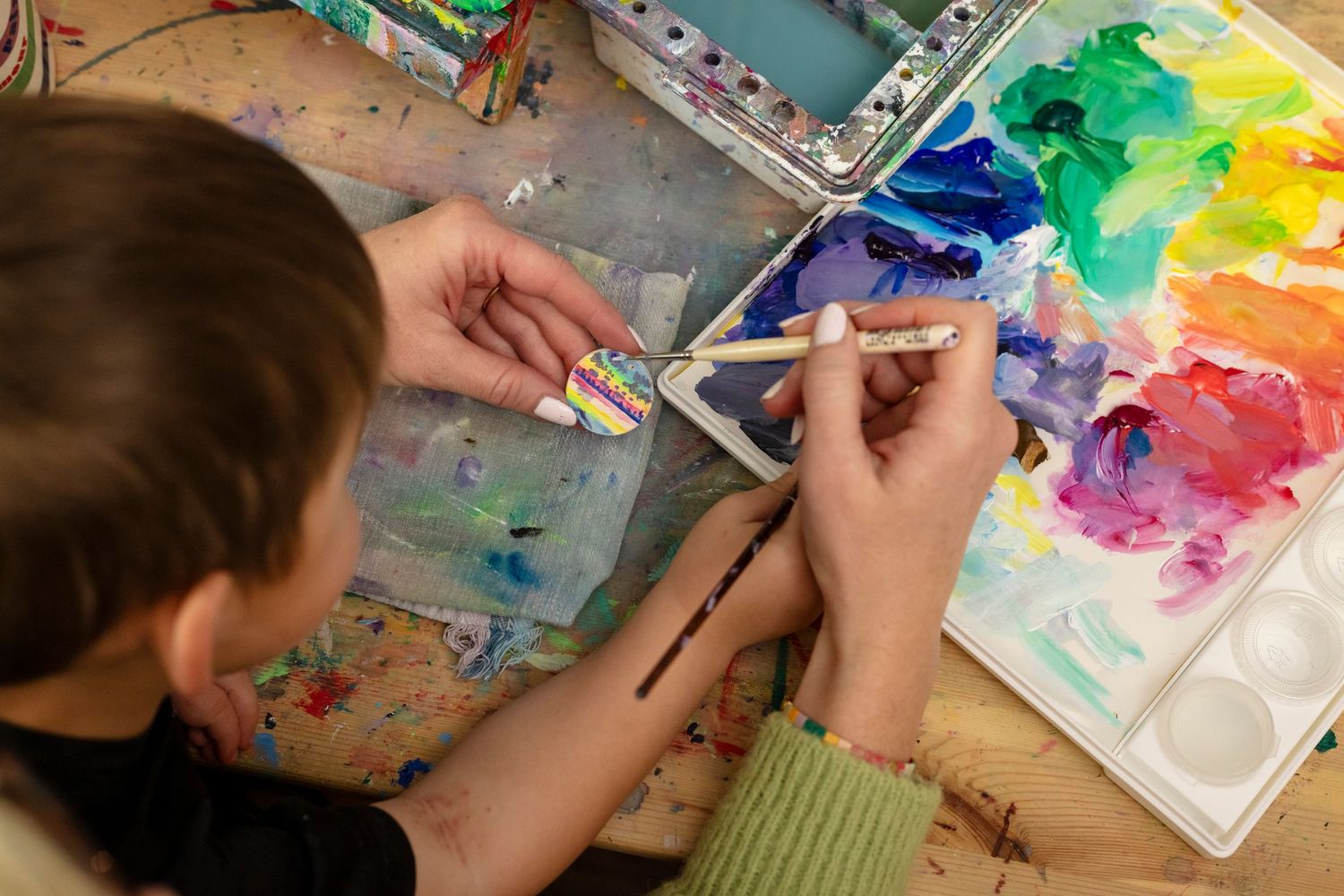 Overhead view of artist using watercolor paints with a small child and brushes with a vibrant rainbow palette on display.