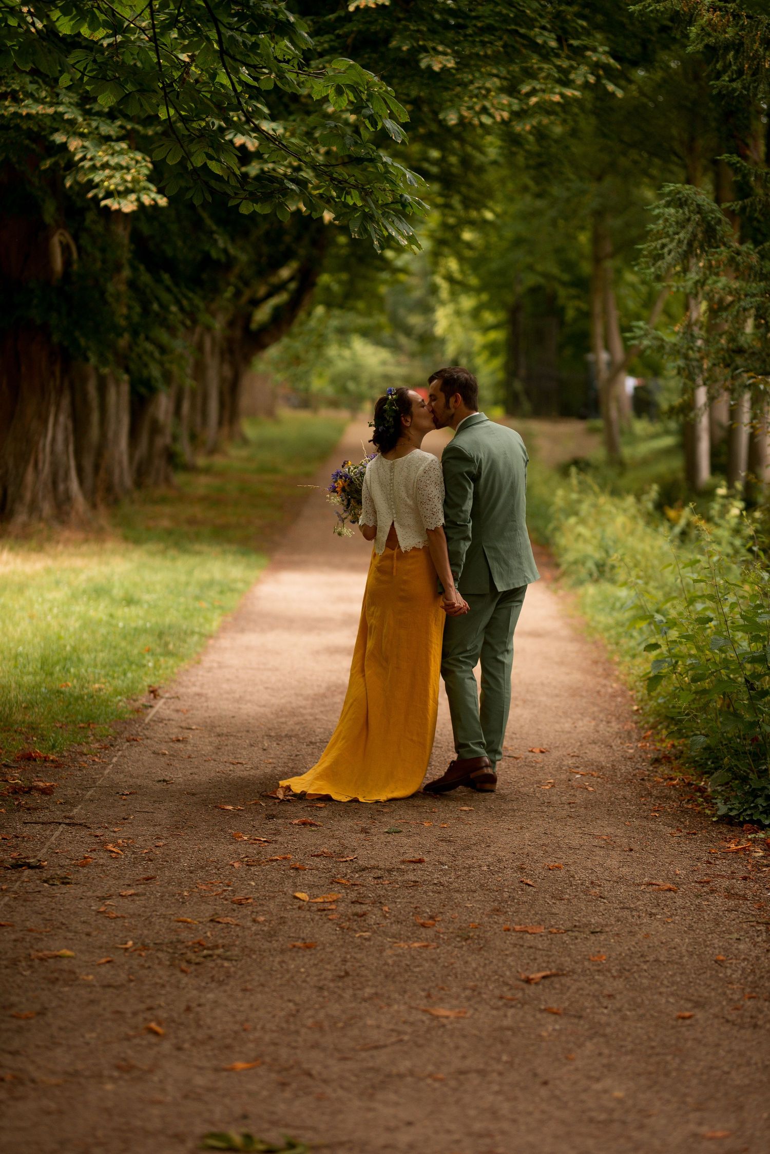 bride wearing white and yellow holding a wild boquet kisses her groom as they walk through a tree lined path