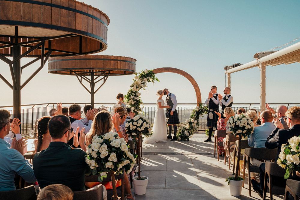 Outdoor rooftop wedding ceremony with wooden arches and guests seated beneath a blue sky.