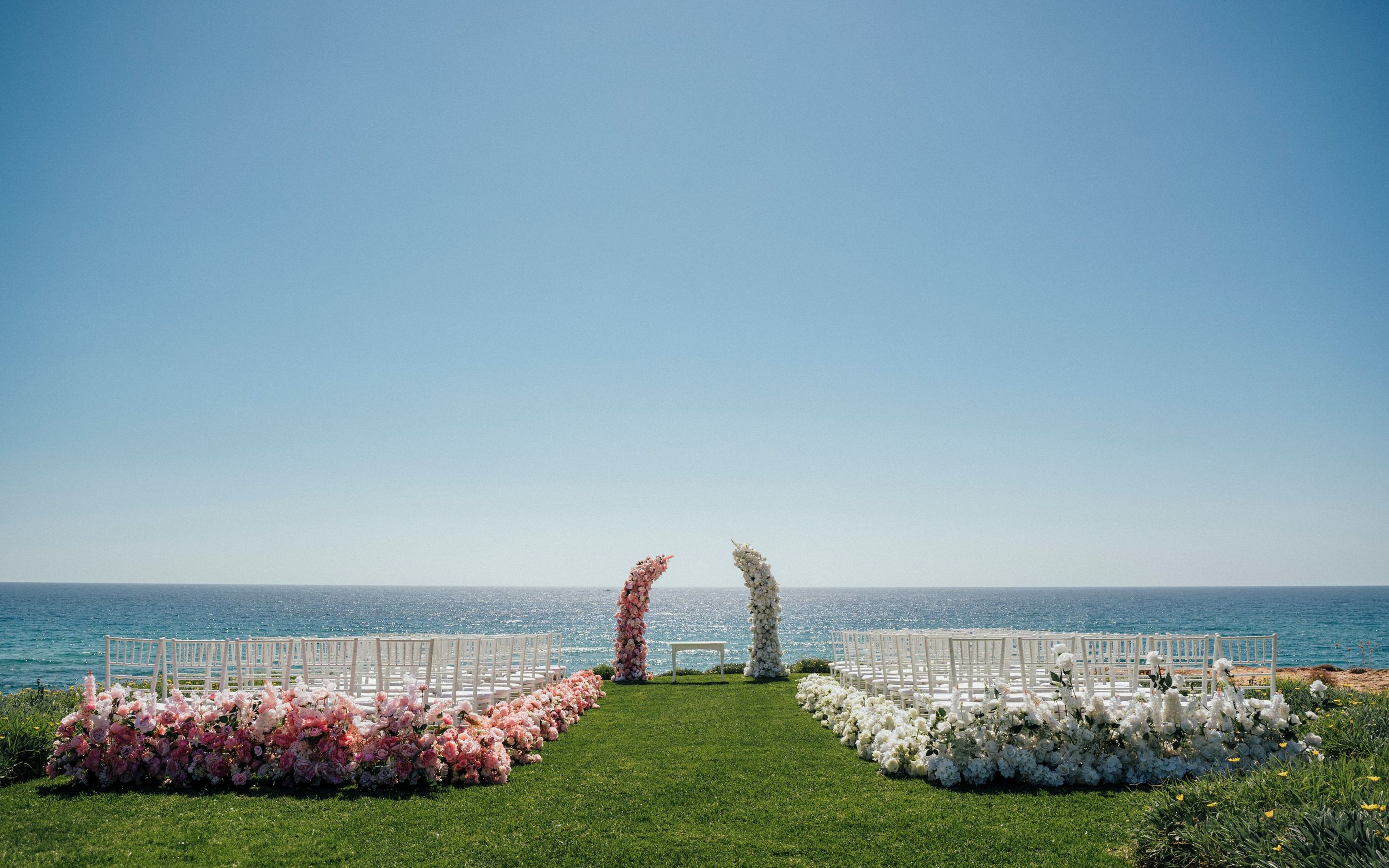Oceanfront wedding ceremony setup with white chairs divided by a grass aisle lined with pink flowers on a sunny day.