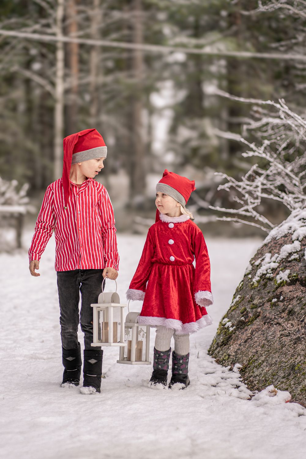 Children carrying lanterns while walking through snowy forest in matching red holiday outfits.