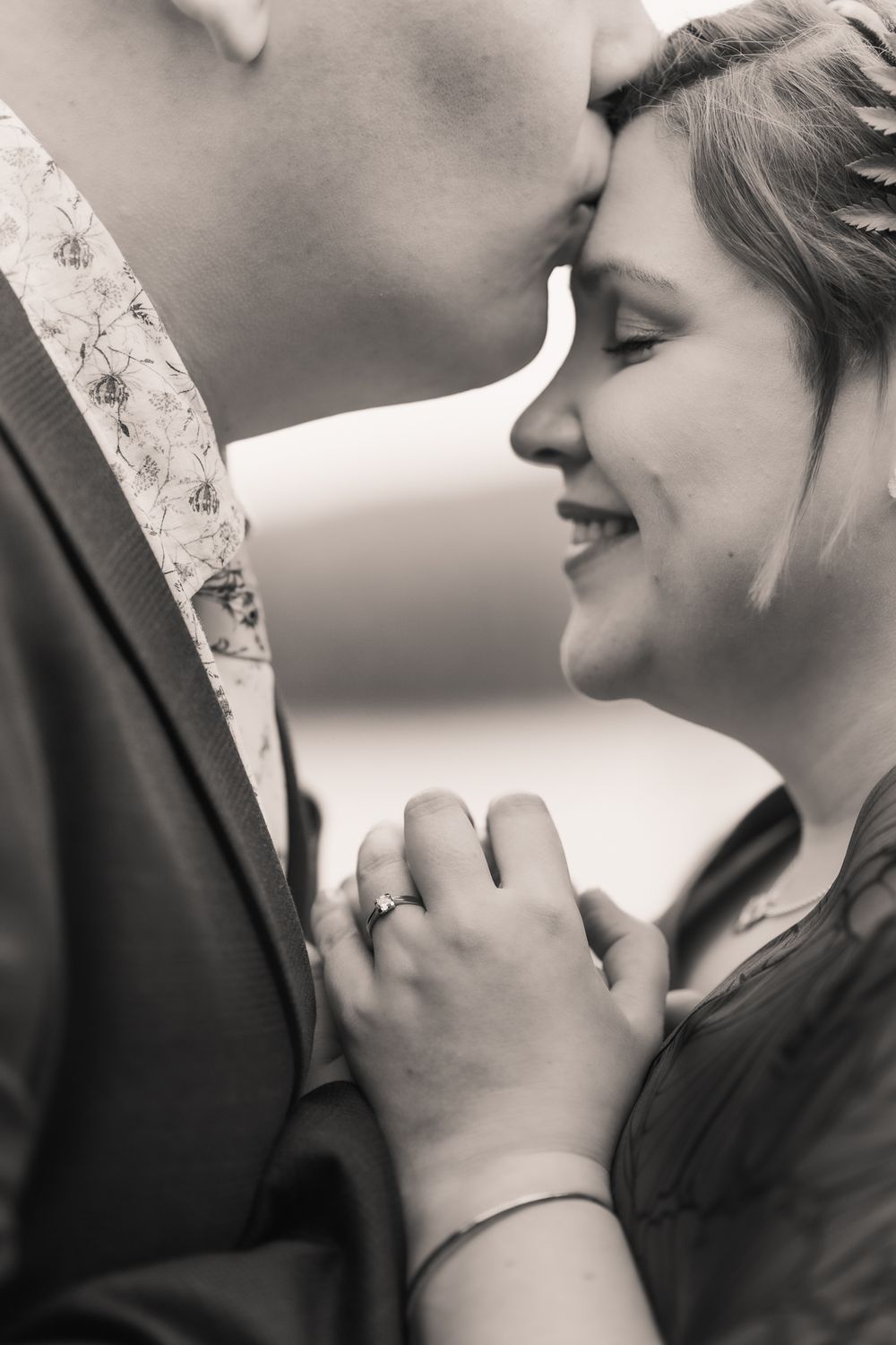 Black and white close-up of a romantic moment showing an affectionate kiss and smile with an engagement ring visible.