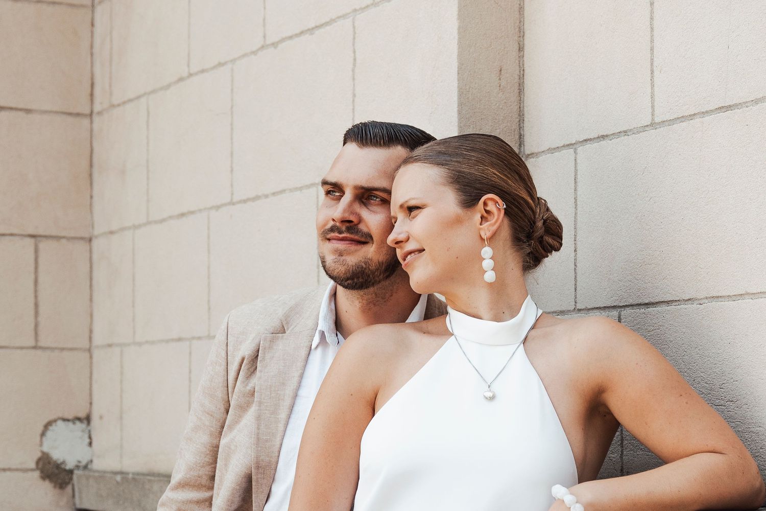 Stylish couple in formal attire pose against neutral wall during wedding photoshoot.
