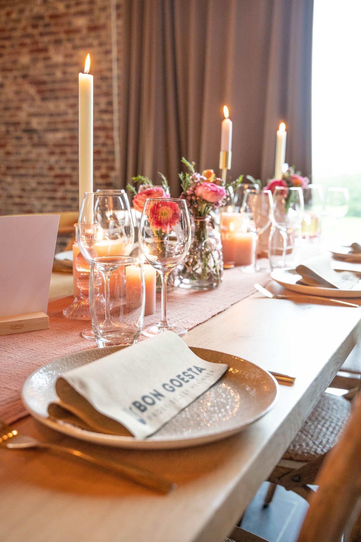 Elegant table setting with tall white candles, pink flowers, and wine glasses against exposed brick wall.